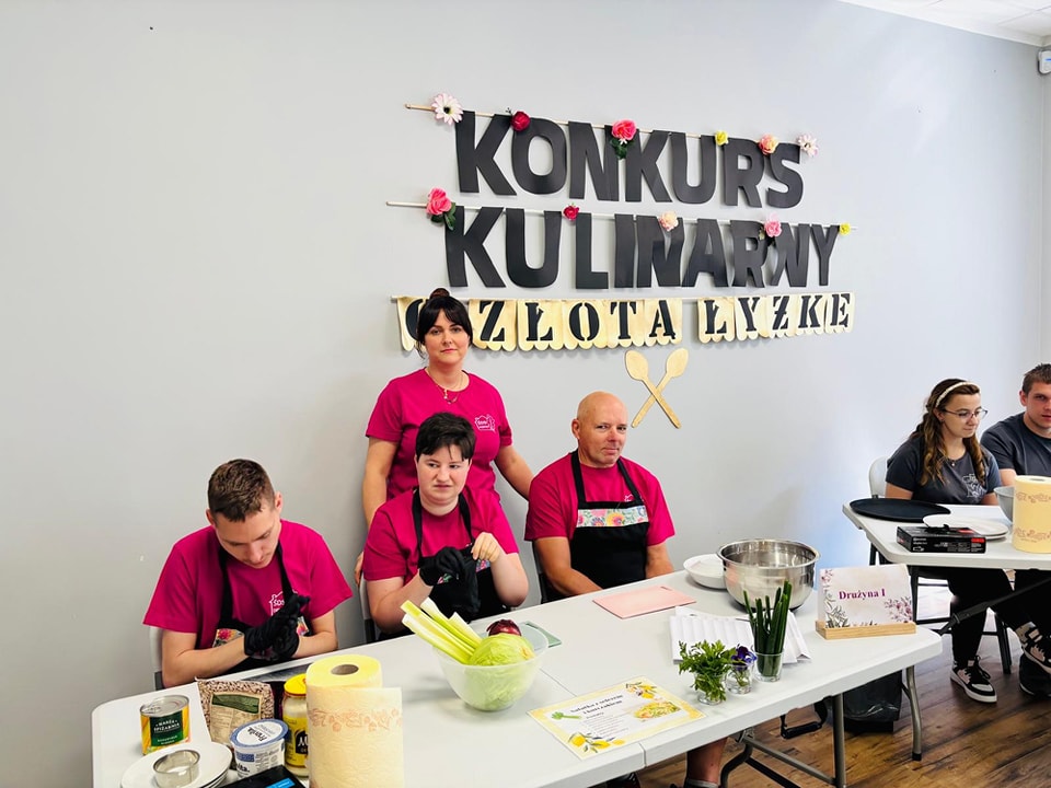 Drużyna w różowych koszulkachsiedzi przy stole, na którym są miski i inne rzeczy do przygoowania potrawy. Na ścianie napis  konkurs kulinarny o Złotą Łyżkę