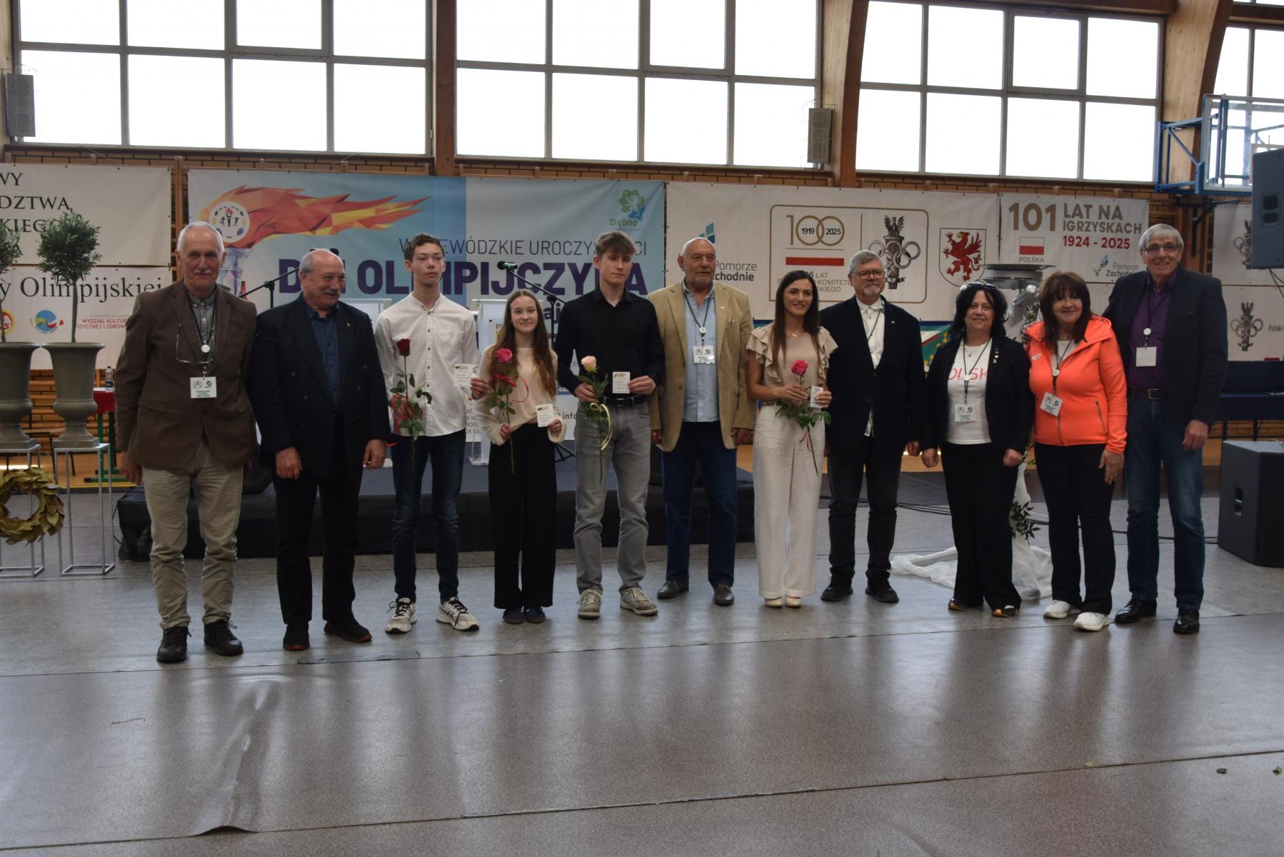 Wyrózneini uczniowie kółkami olimpijskimi wraz z olimpijczyakami stoja w rzędzie na hali