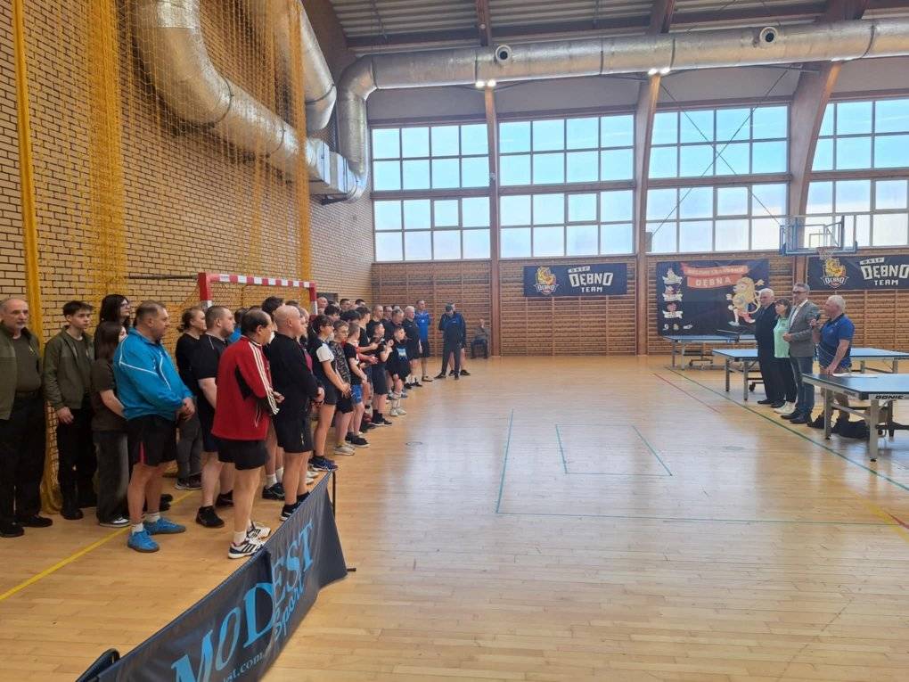 Na hali sportowej stoi grupa ludzi. Po prawe organizatorzy i burmistrz Debna, po lewej zawodnicy