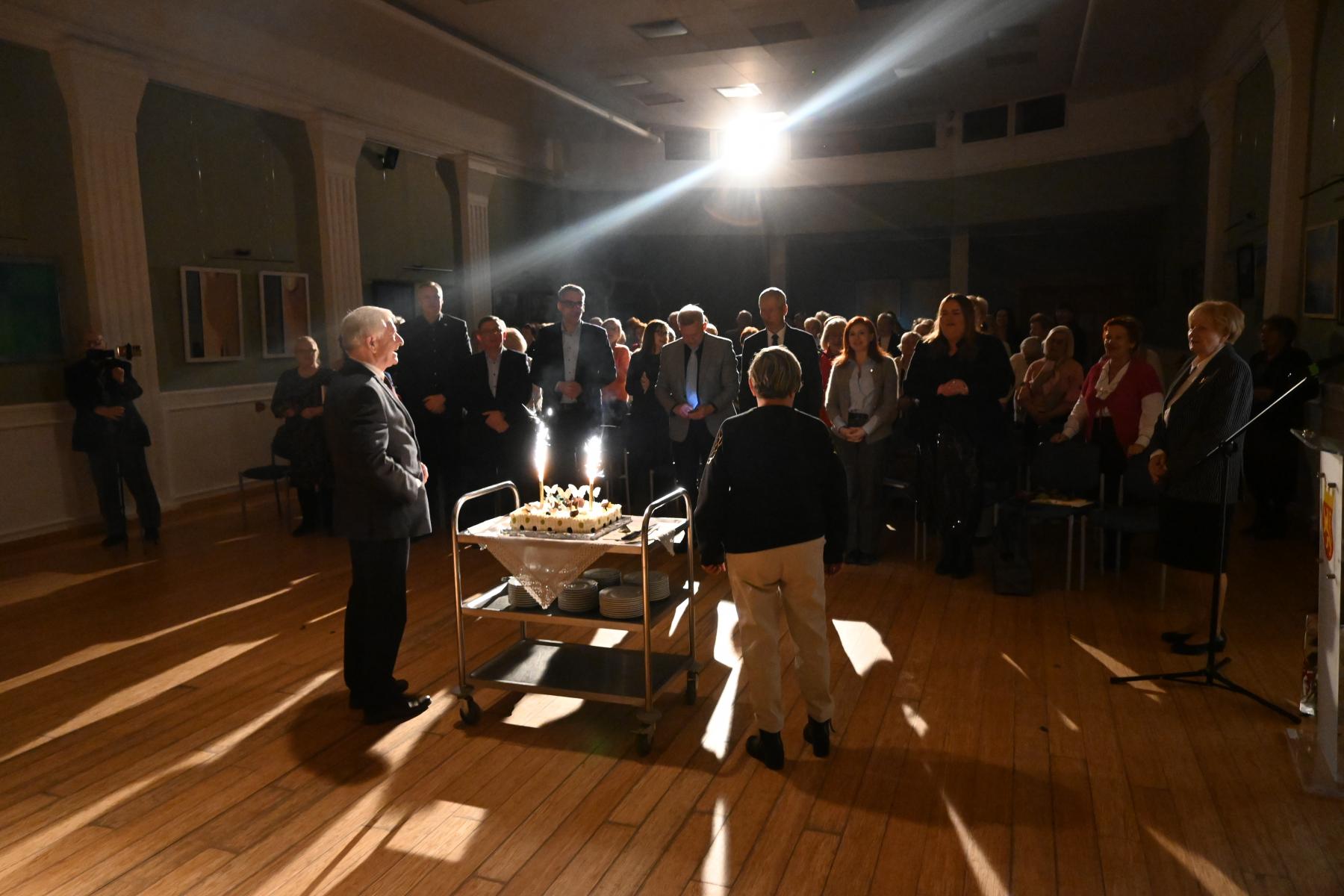 ludzie stoją w przyciemnionej sali. Na środku stoi stolik z tortem