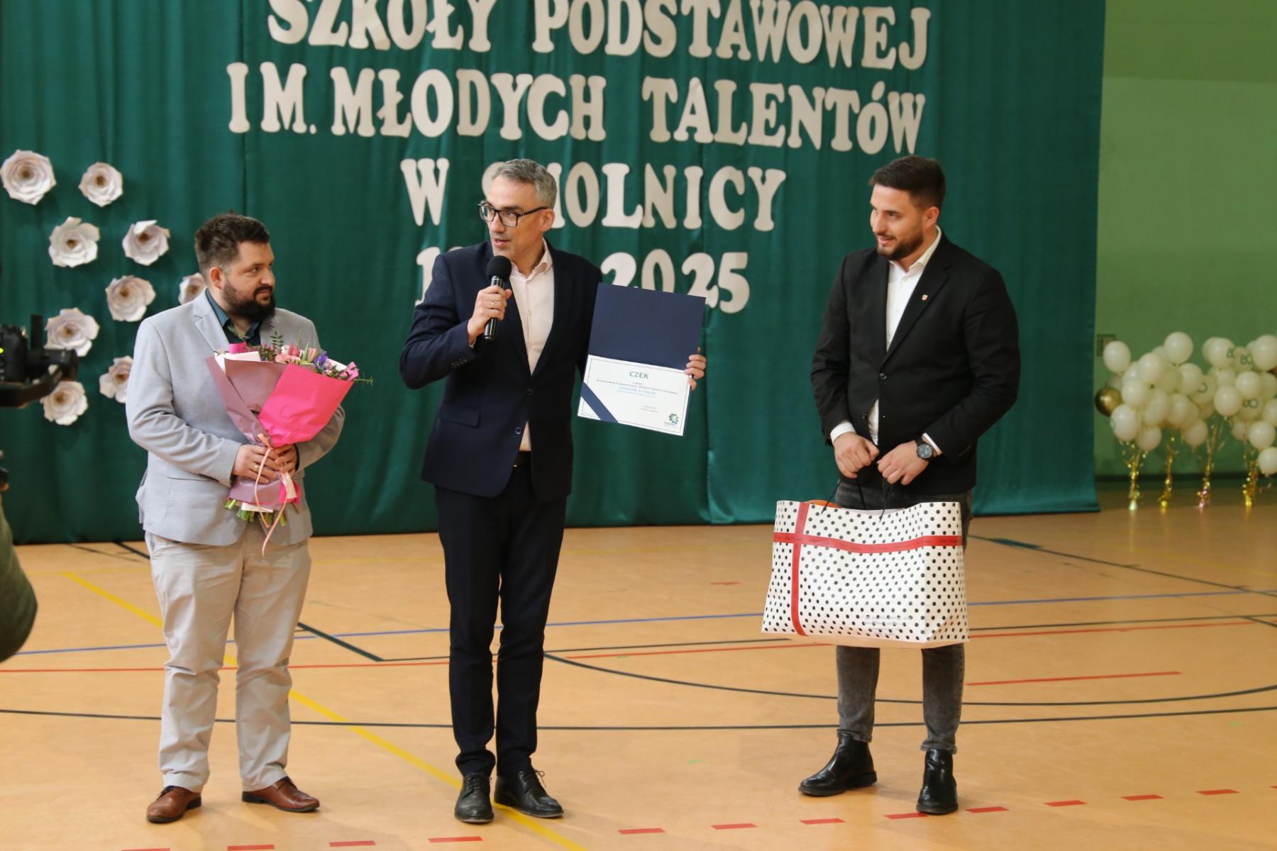 od lewej, radny, burmistrz i wiceprzewodniczący rady, burmistrz z mikrofonem przemawia, radni trzymają kwiaty i prezent w torebce