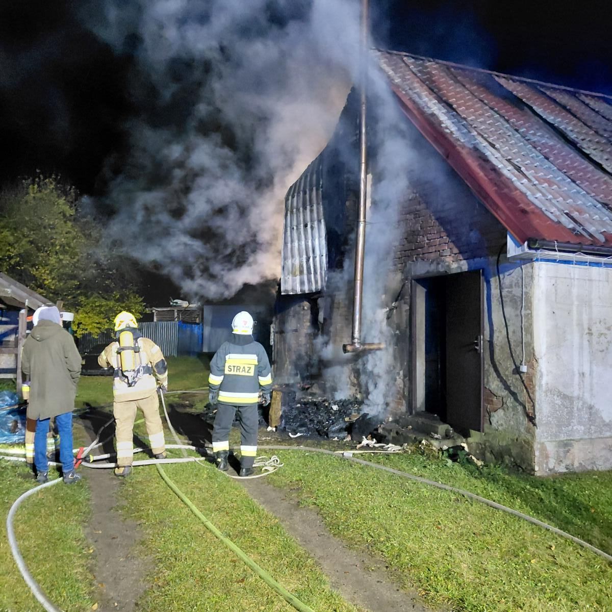 spalony budynek nocą. Tyłem, przed budynkeim stoją strażacy i mężczyzna 