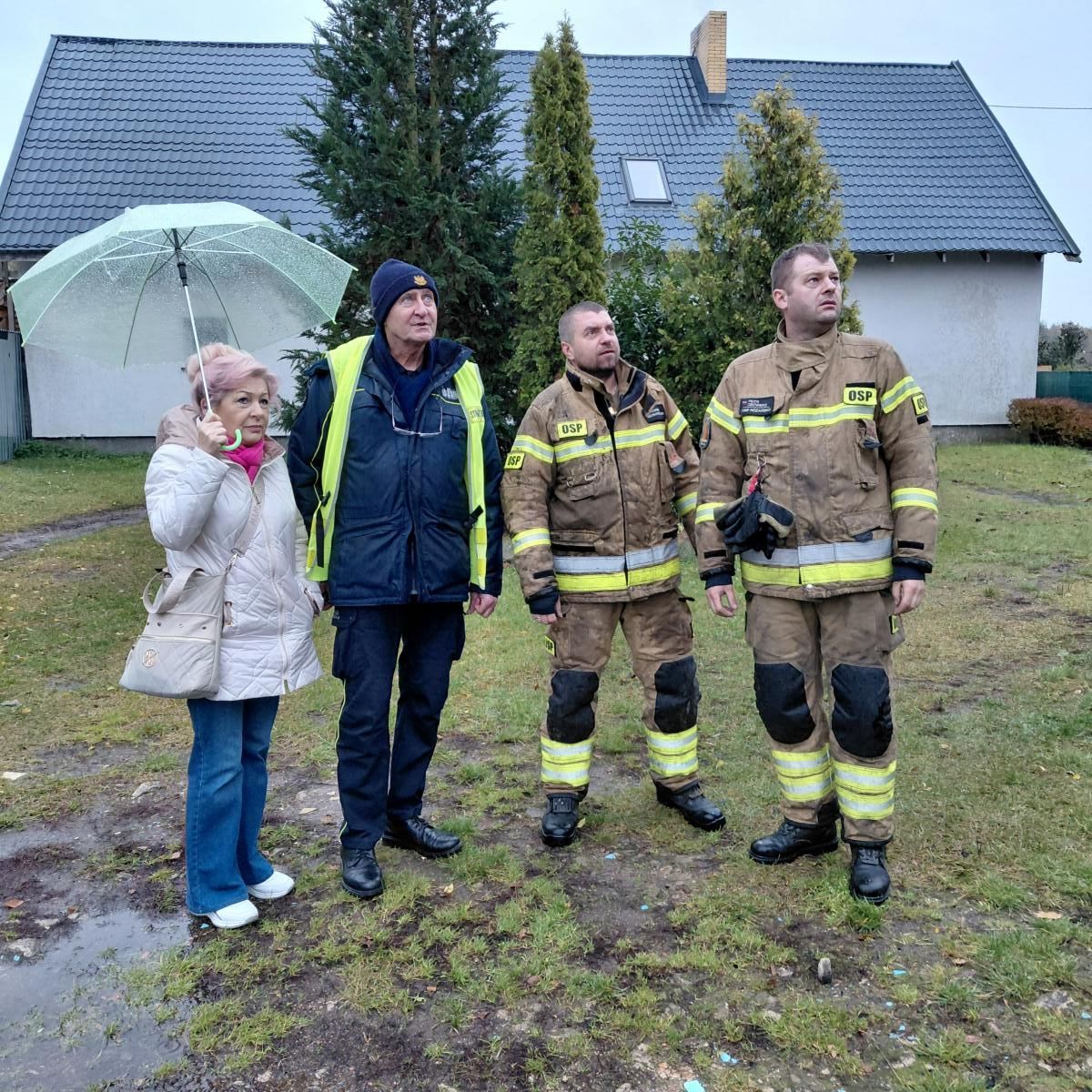 Od lewej w dzień na dworze stoją: kobieta, komendant straży miejskiej, dwóch strazaków w mundurach, w tle dach domu
