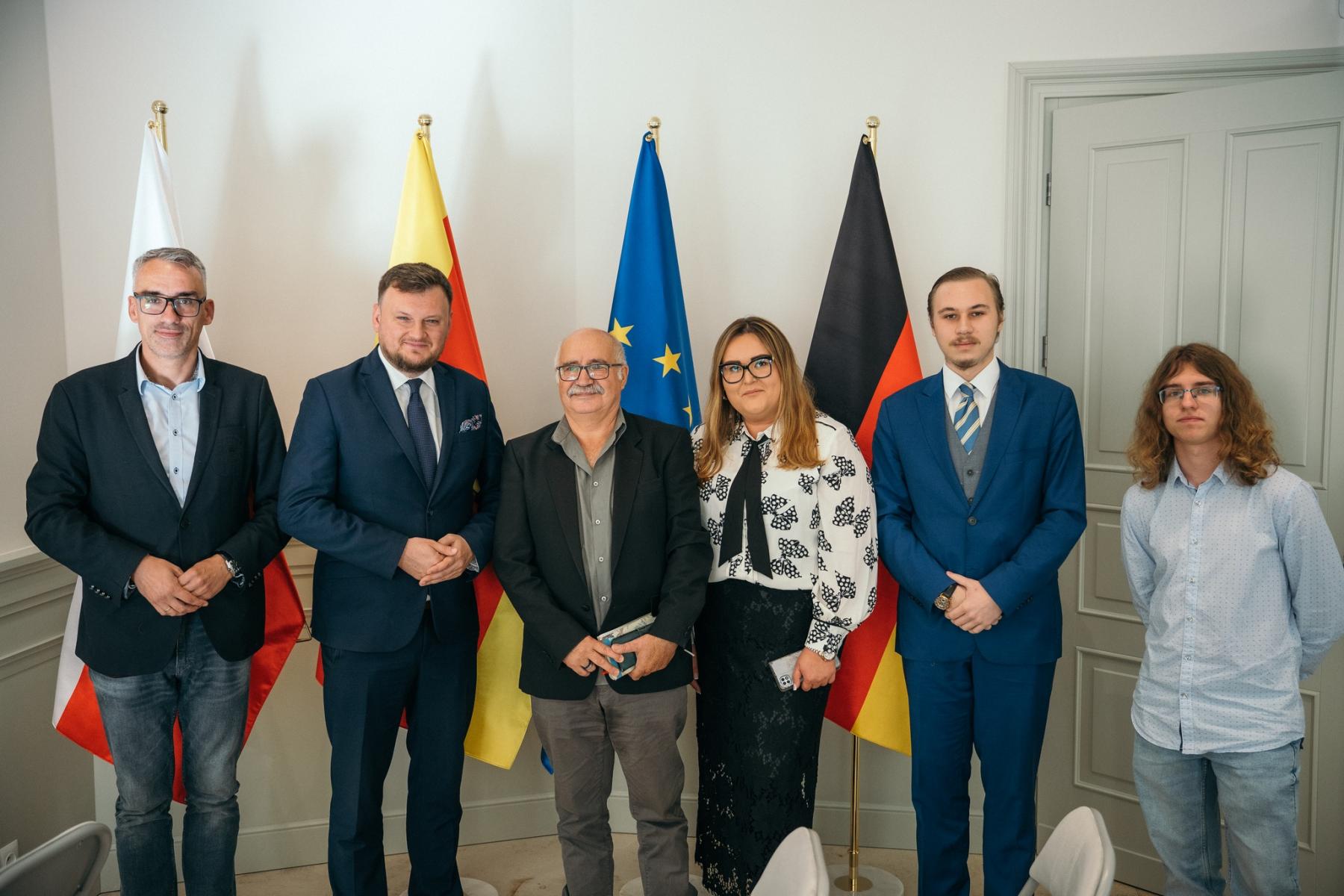 Od lewej na tle flaga stoją: burmistrz Debna, dwóch mężczyzn, kobieta, dwóch młodych mężczyzn