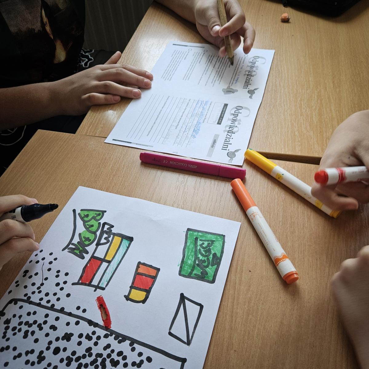 dziecięca ręka trzyma ołówek, na blacie leży zadrukowana kartka. Obok kartka od bloku z kolorowymi rysunkami, pisaki kolorowe