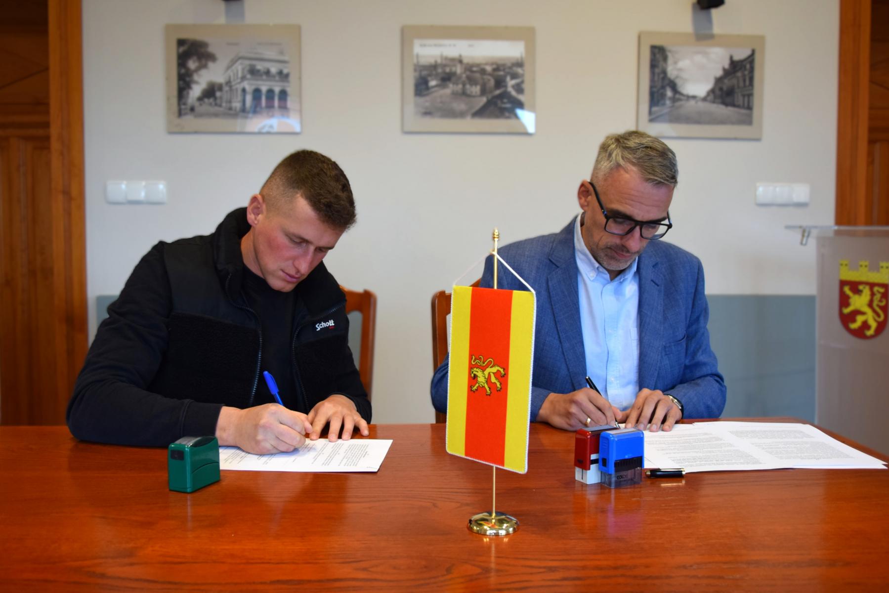 Dwóch mężczyzn siedzi przy stole w sali, podpisują dokumenty. w tle na ścianie wiszą obrazki