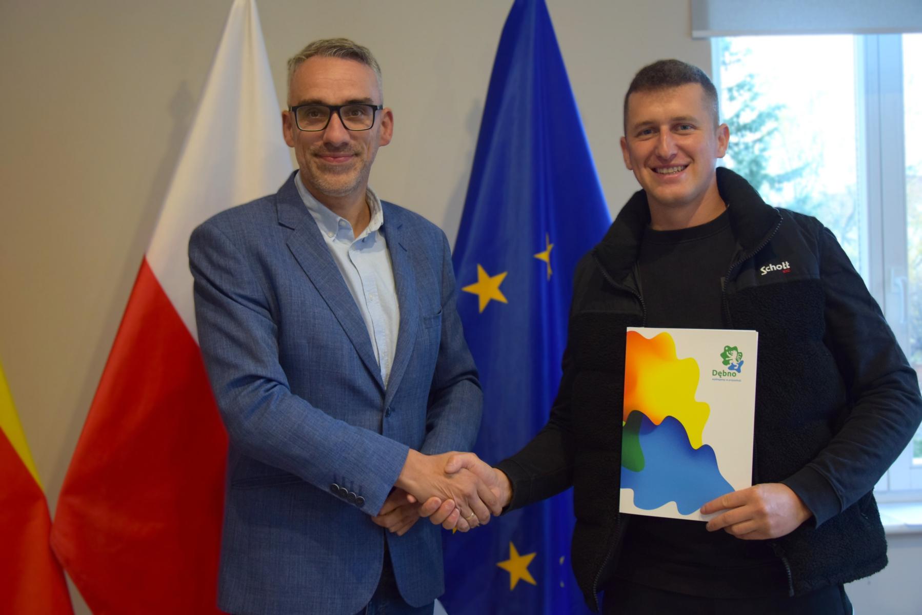 Dwóch mężczyzn stoi w sali,, ściskają sobie dłonie. Od lewej burmistrz, prezes klubu, w tle flaga 