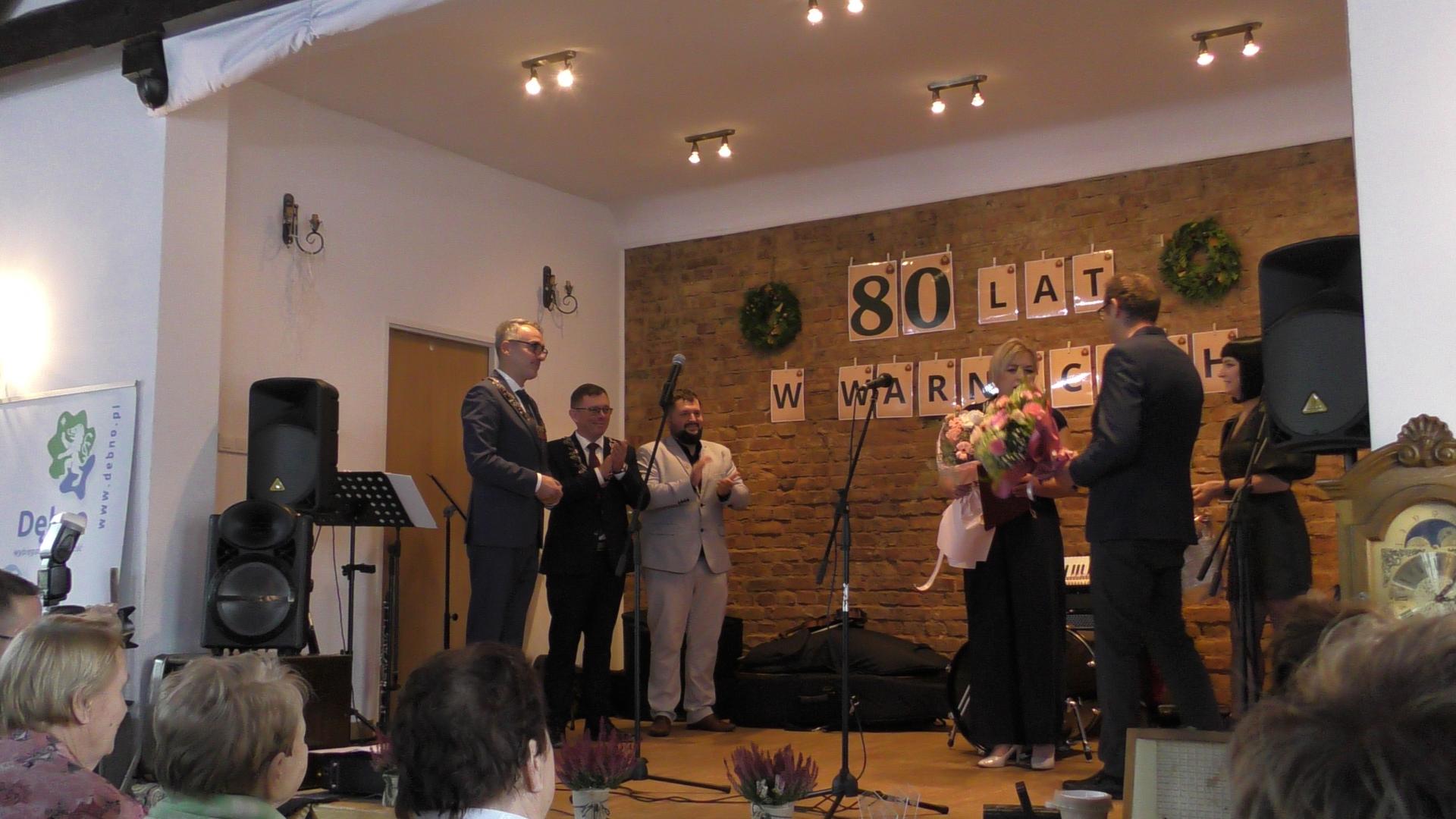 Na scenie stoją przedstwaieicele władzy (po lewej stronie stoi trzech mężczyzn) po prawej wyr&oacute;zniona medalem z kwiatami i mężczyzna i kobieta