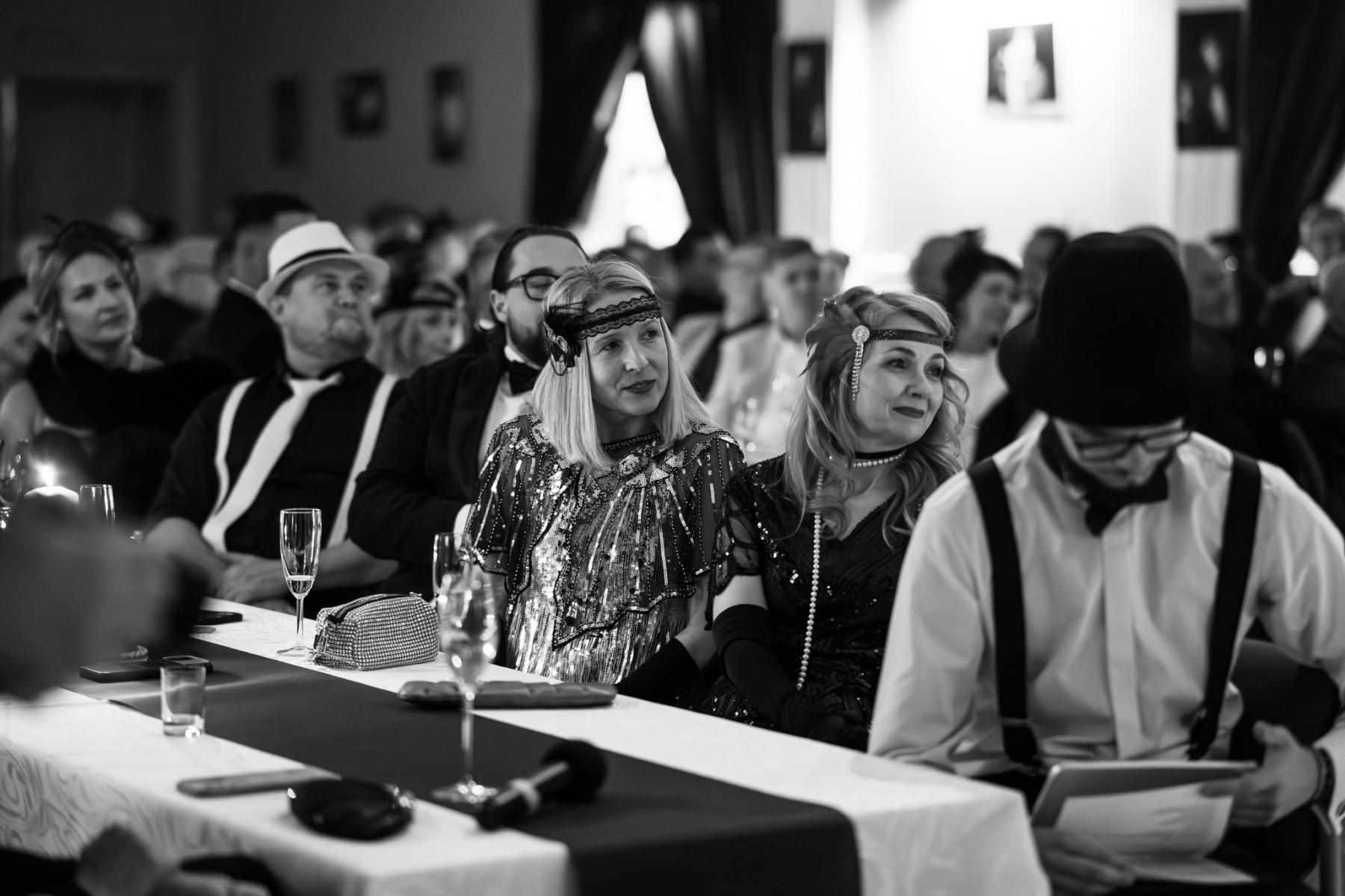 Kobiety i mężczyzni siedzą przy stole w strojach stylizowanych na lata 20, zdjęcie czarno-białe