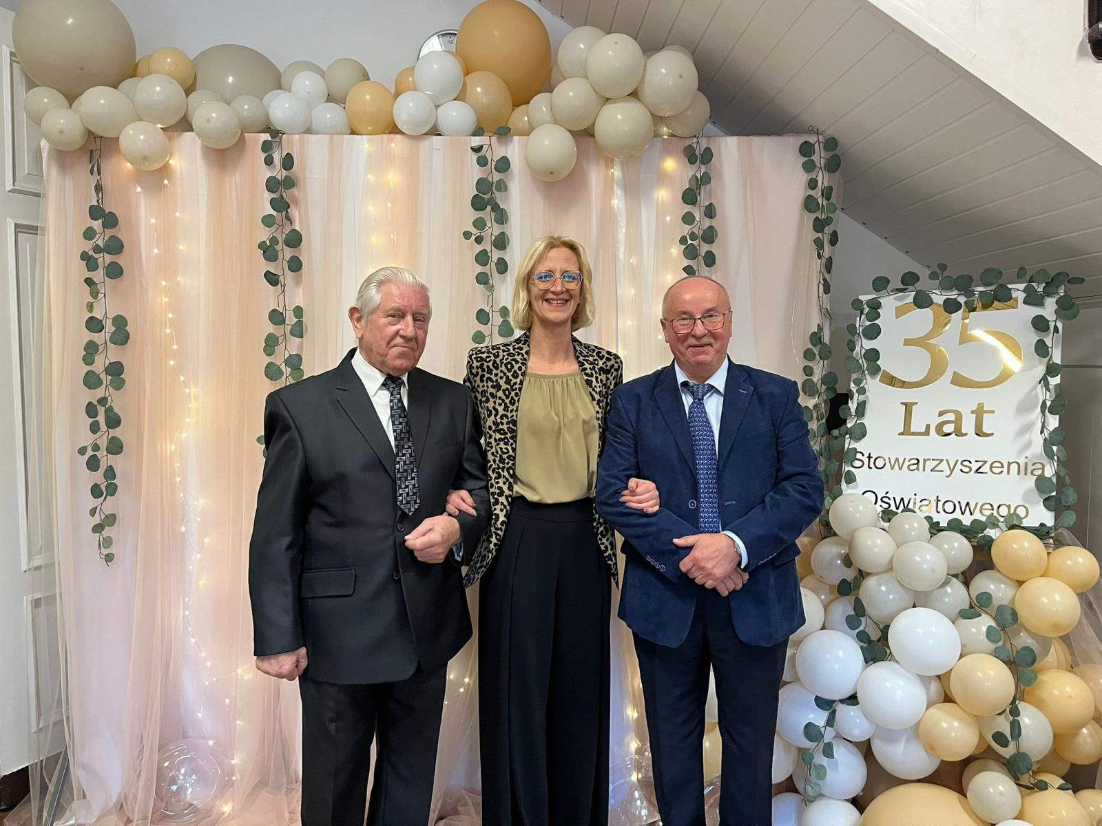 Dwóch mężczyzn i kobieta w środku stoja na tle ścianki z balonami 