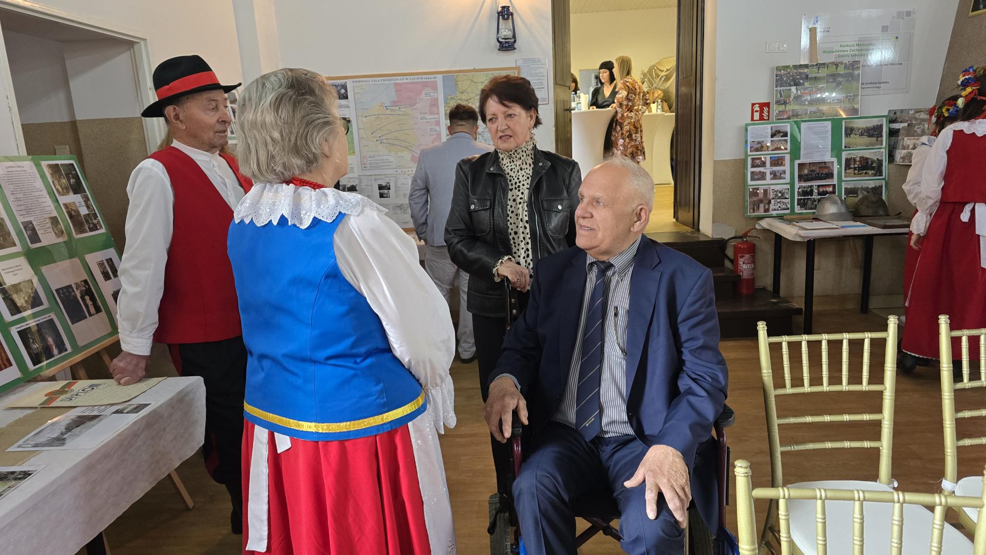 Na w&oacute;zku siedzi mężczyzna, za nim stoi kobieta, obok w stroju ludowym stoją kobieta i mężczyzna 