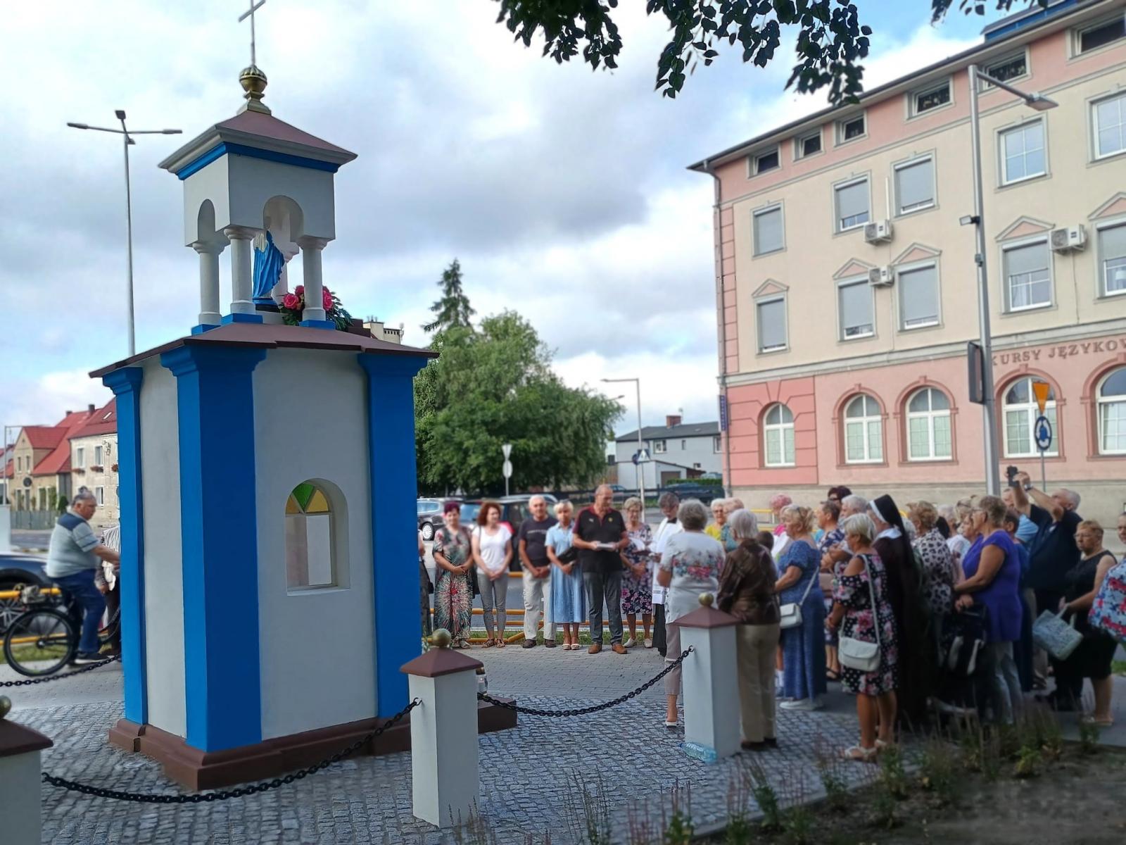 Niebiesko-biała kapliczka odnowiona. z przodu stoi grupa ludzi i ksiądz. w tle budynek, niebieskie niebo z chmurami