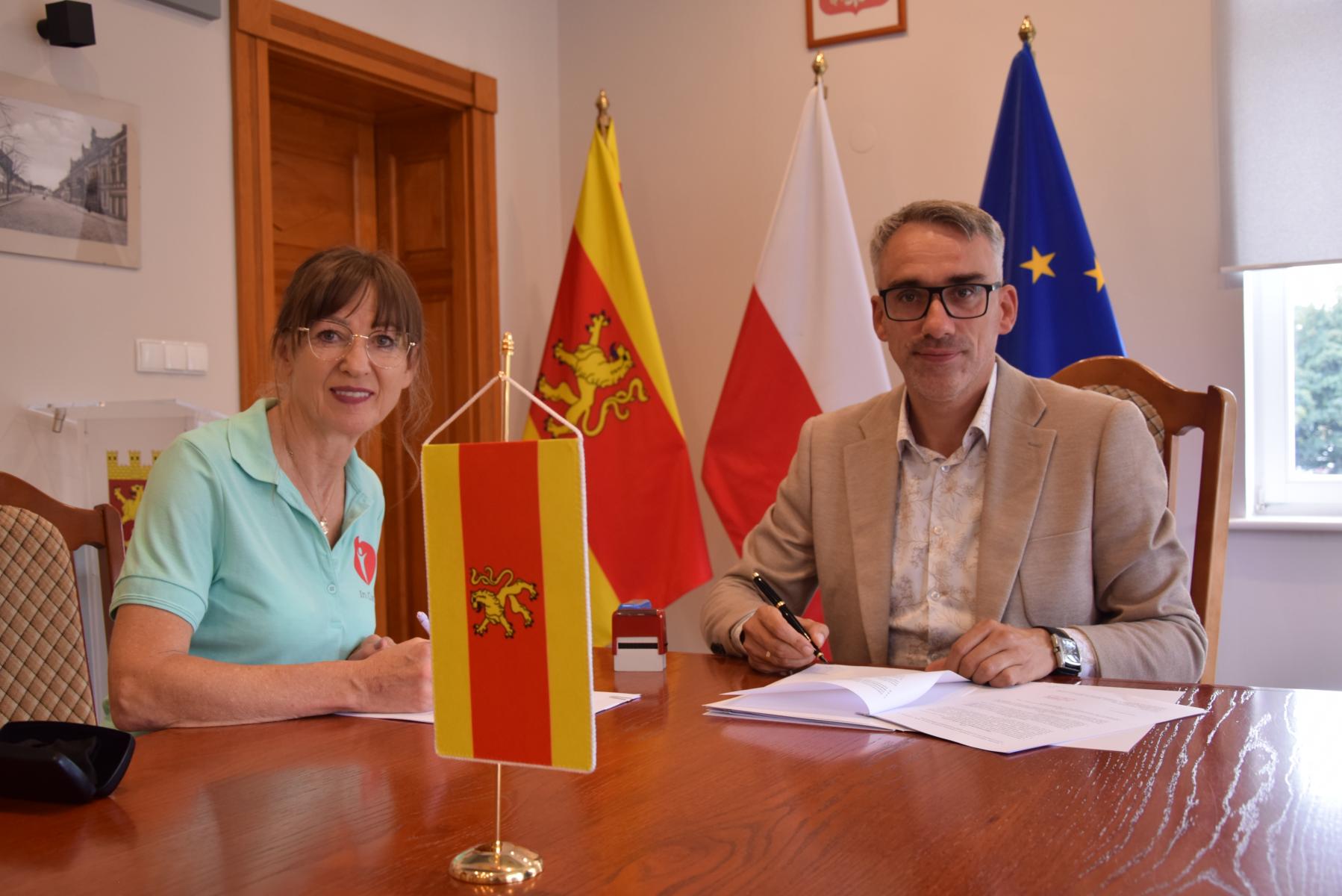 Przy stole, w sali siedzą pani Aneta i burmistrz. W tle flagi. Na stole leżą dokumenty 