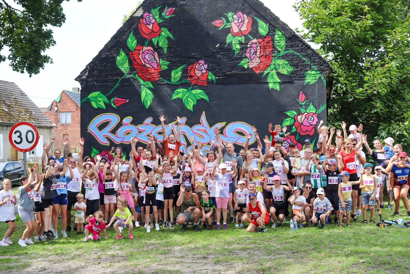 Duża grupa ludzi stoi, ludzie mają podniesione ręce, na koszulkach numery startowe. W tle ściana n=budynku z muralem z różami i napis Różańsko