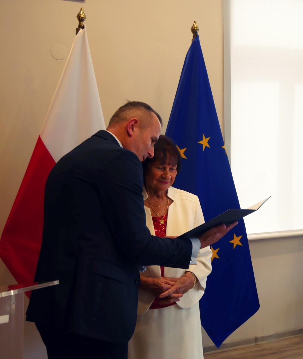 Mężczyzna (M. Łapecinski) wręcza dyplom pani Anieli, w tle dlaga Polski i Unii Europejskiej