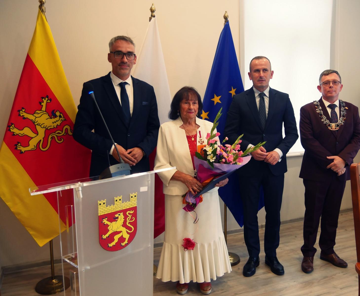 Od lewej, w sali stoją burmistrz Dębna W. Czepułkowski, Aniela Głodna z kwiatami w dłoniach, Członek Zarządu M. Łapeciński, Przewodniczący Rady Miejskiej J. Mazurowski