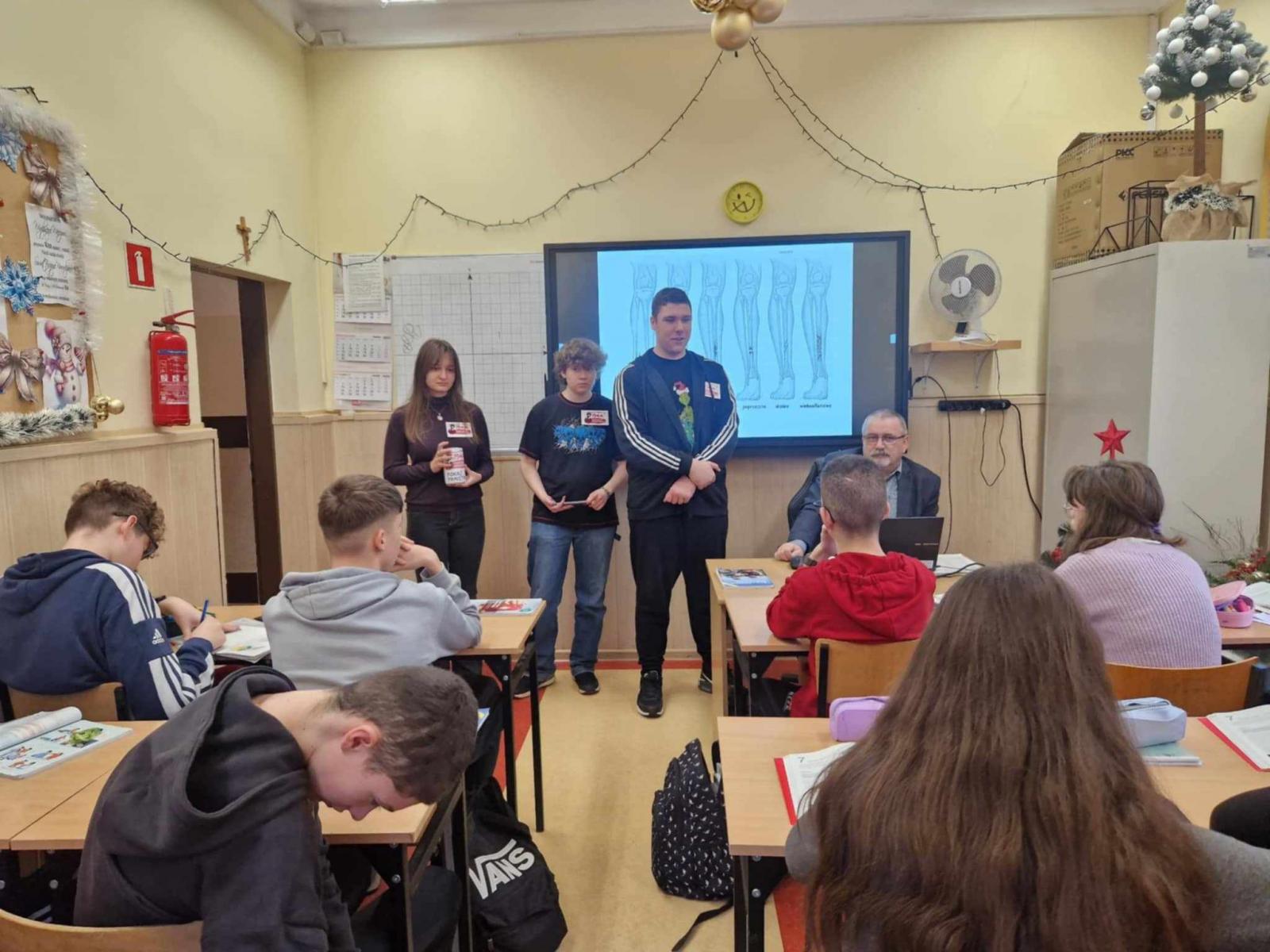 Trzech uczniów stoi przodem do reszty uczniów, którzy siedzą w ławkach. 