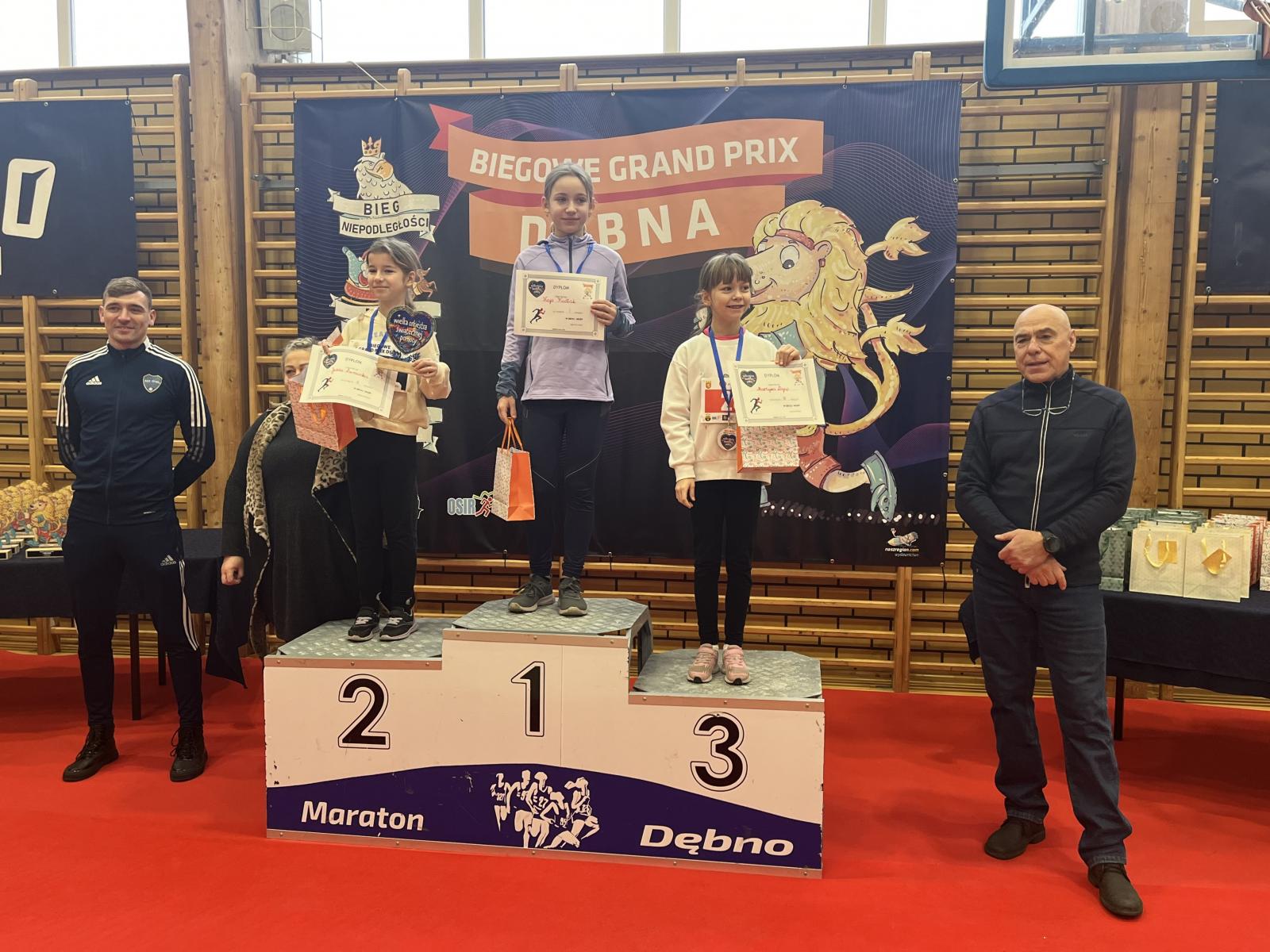 Trzy dziewczynki stoją na podium, trzymają dyplomy i torebki w rękach. obok po lewe i po prawej stoją mężczyzni