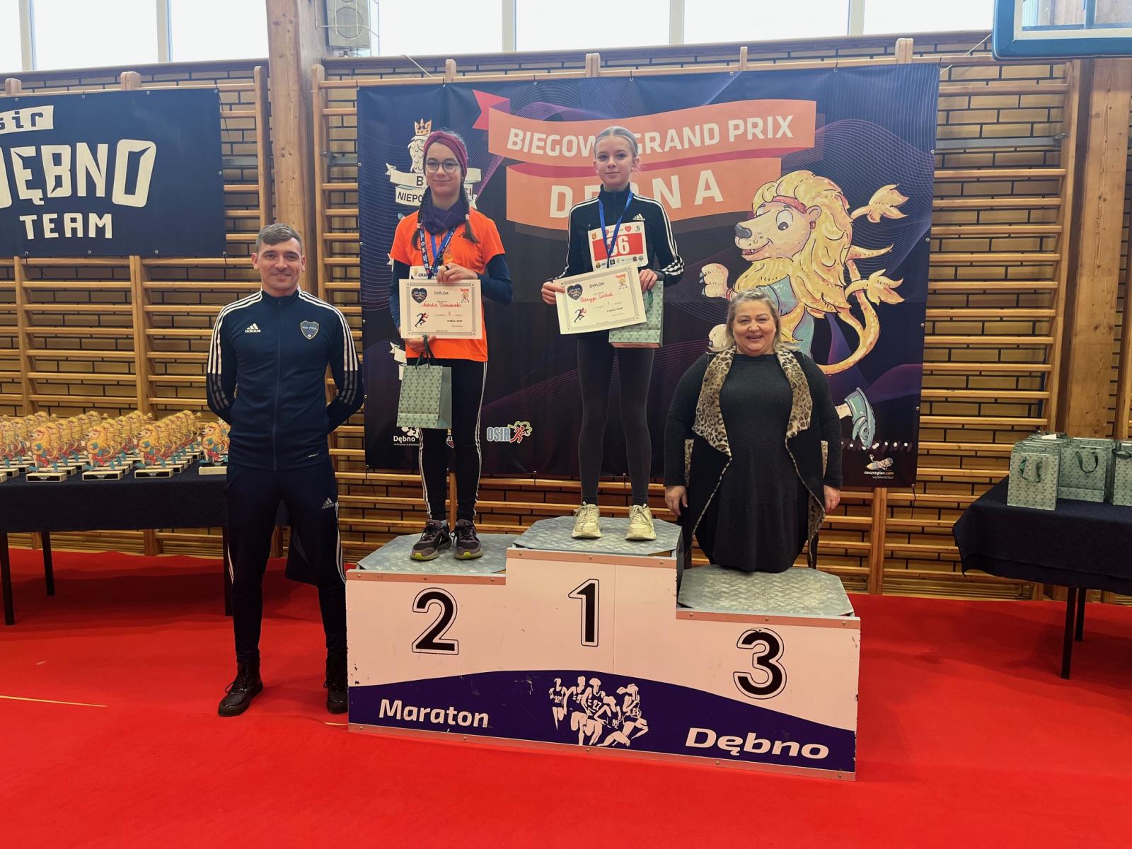 Dwie dziewczynki stoją na podium, trzymają dyplomy i torebki w rękach. obok po lewej stoi mężczyzna, po prawej kobieta