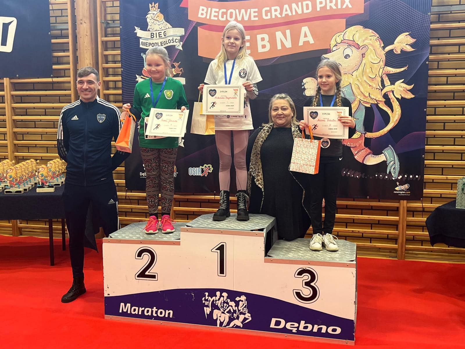 Trzy dziewczynki stoją na podium, trzymają dyplomy i torebki w rękach. obok po lewej stoi mężczyzna 