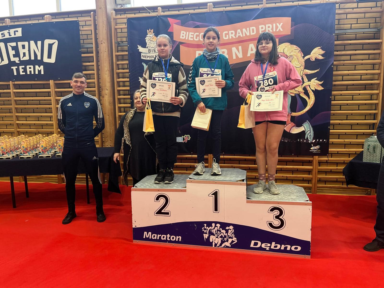 Trzy dziewczynki stoją na podium, trzymają dyplomy i torebki w rękach. obok po lewej stoi mężczyzna 