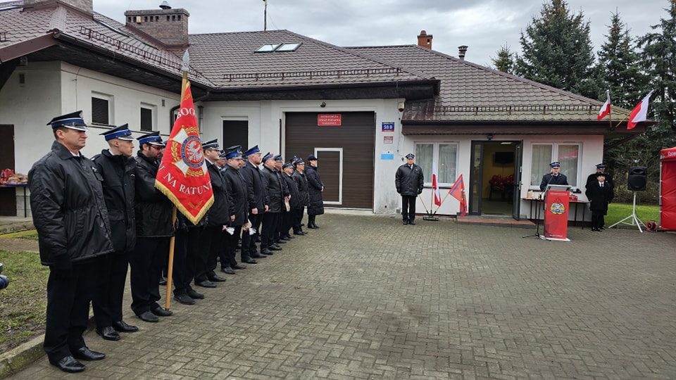 Strażacy stoją w szeregu na placu, przed budynkiem 