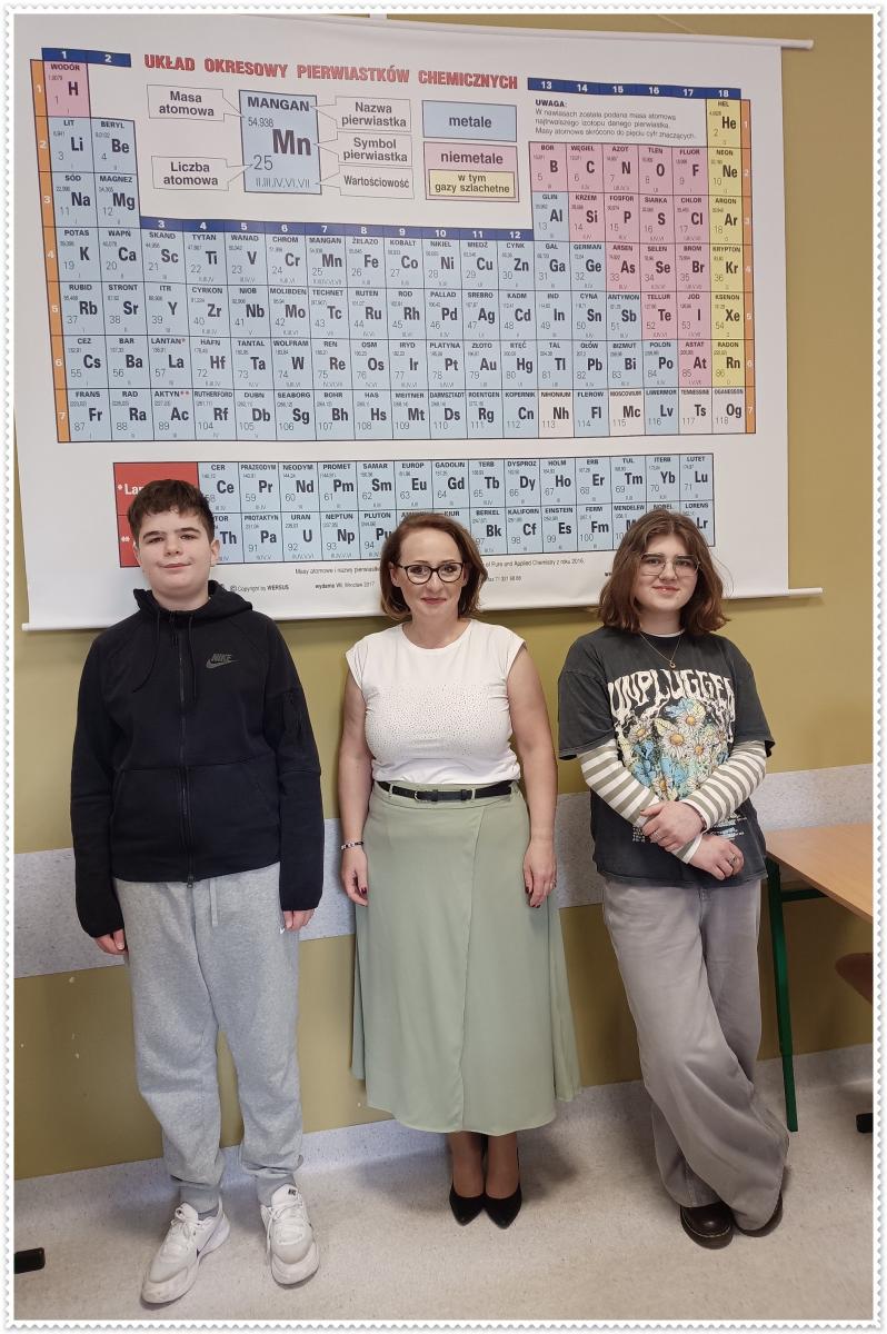 Stoją w klasie uczen, nauczycielka, uczennica. Na ścianie wisi plansza z pierwiastkami