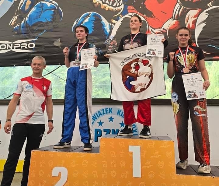 Trzy młode dziewczyny stoją na podium. O lewej stoi mężczyzna, druga na podium Aleksandra