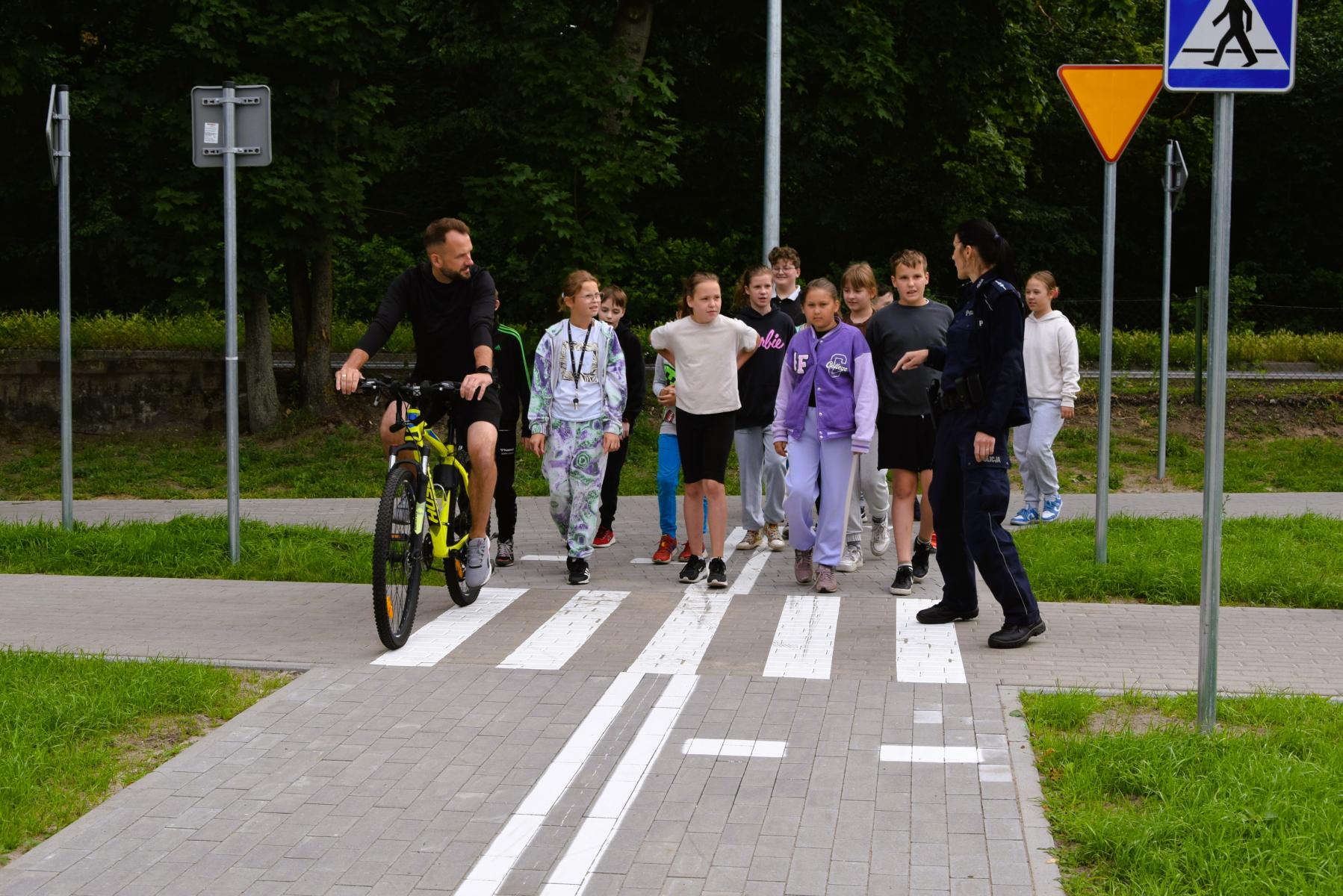 Uczniowie idą z policjantką po miasteczku, obok jedzie nauczyciel na rowerze.  