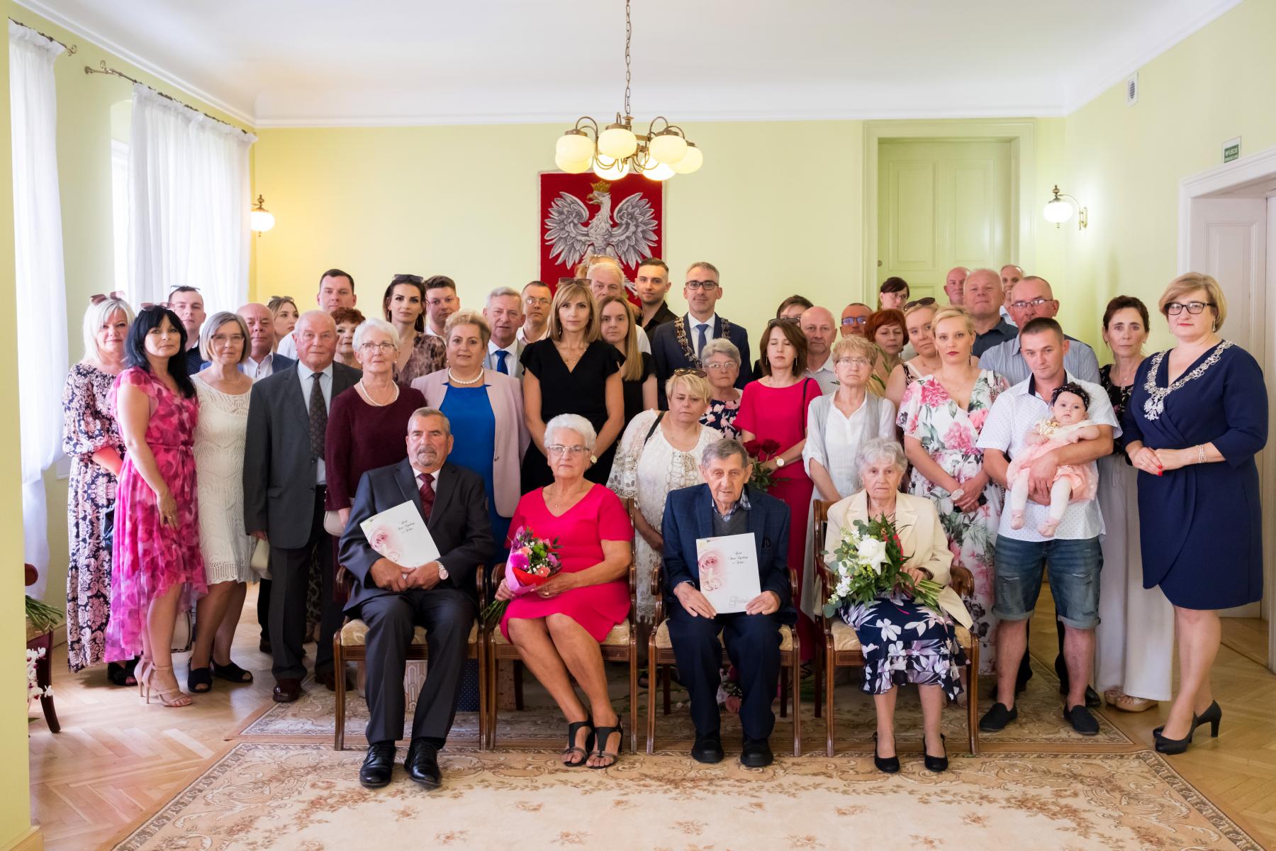 Powiększ zdjęcie. Jubilaci siedzą na krzesełkach, za nimi stoją członkowie rodziny, burmistrz