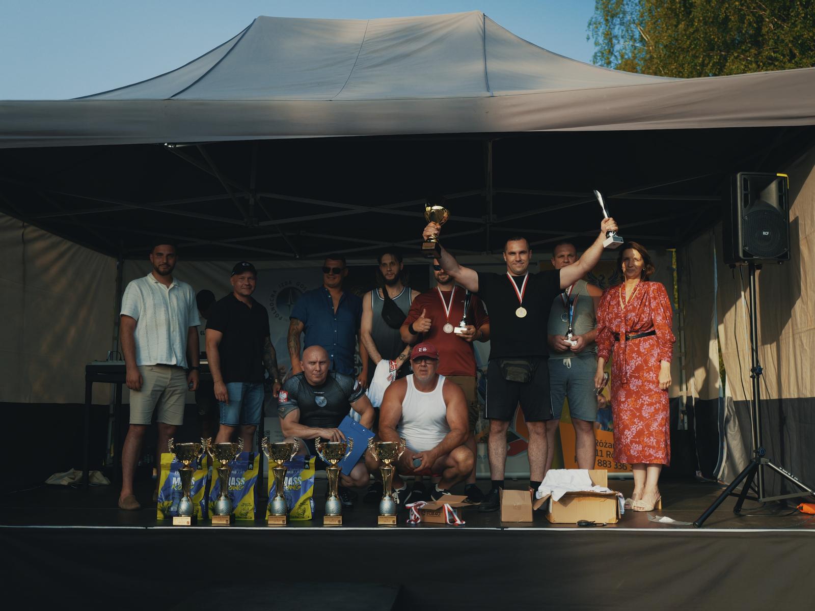 Zawodnicy stoją na scenie. Na przodzie jeden zawodnik trzyma dwa puchary w rękach.