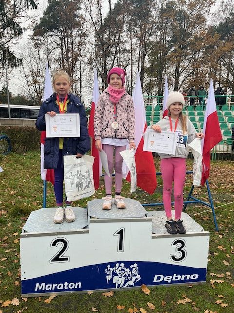 trzy dziewczynki stoją na podium. Z tyłu powiewają biało-czerowne flagi