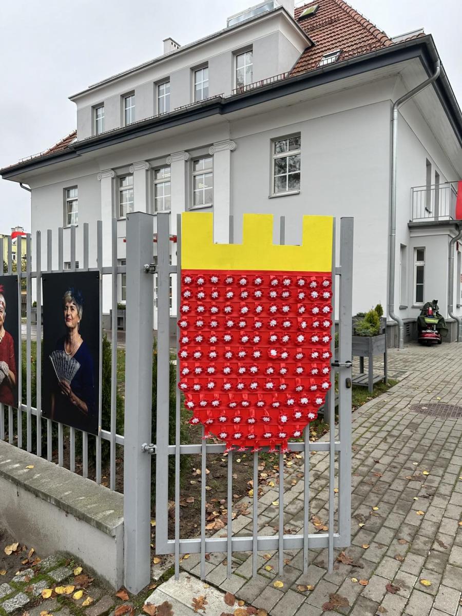 Tarcza w kształcie herbu, na niej przywieszone kokardy, zaieszona na ogrodzeniu bramy, w tle budynek 