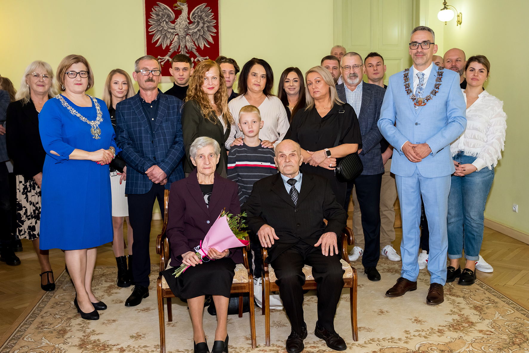 Jubilaci siedzą centralnie na krzesłach, za nimi stoją osoby, po prawej burmistrz Dębna, w niebieskim garniturze 