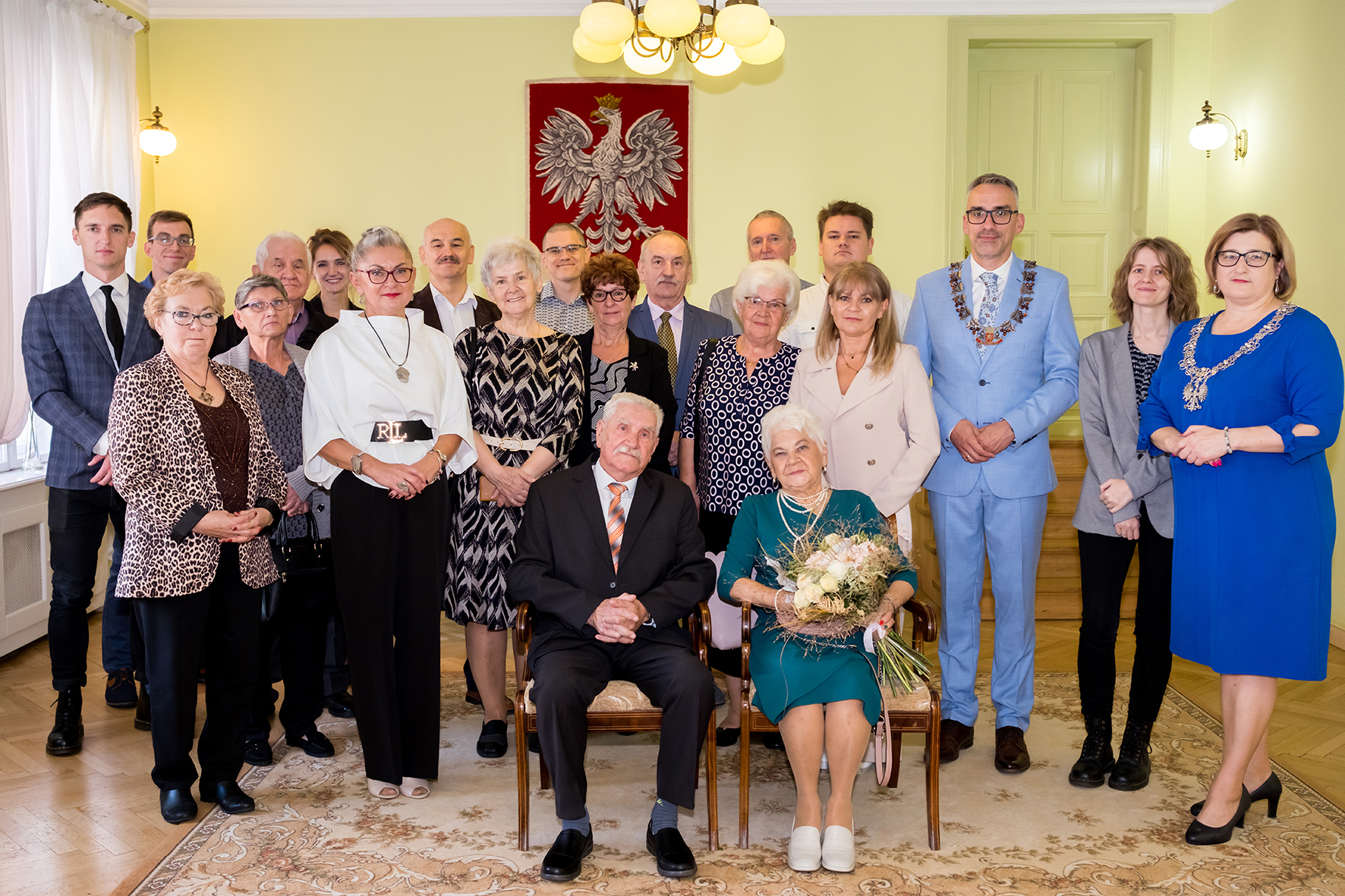Jubilaci siedzą na krzesłach, za nimi stoja osoby z rodziny, w tle na ścianie wisi godło Polski