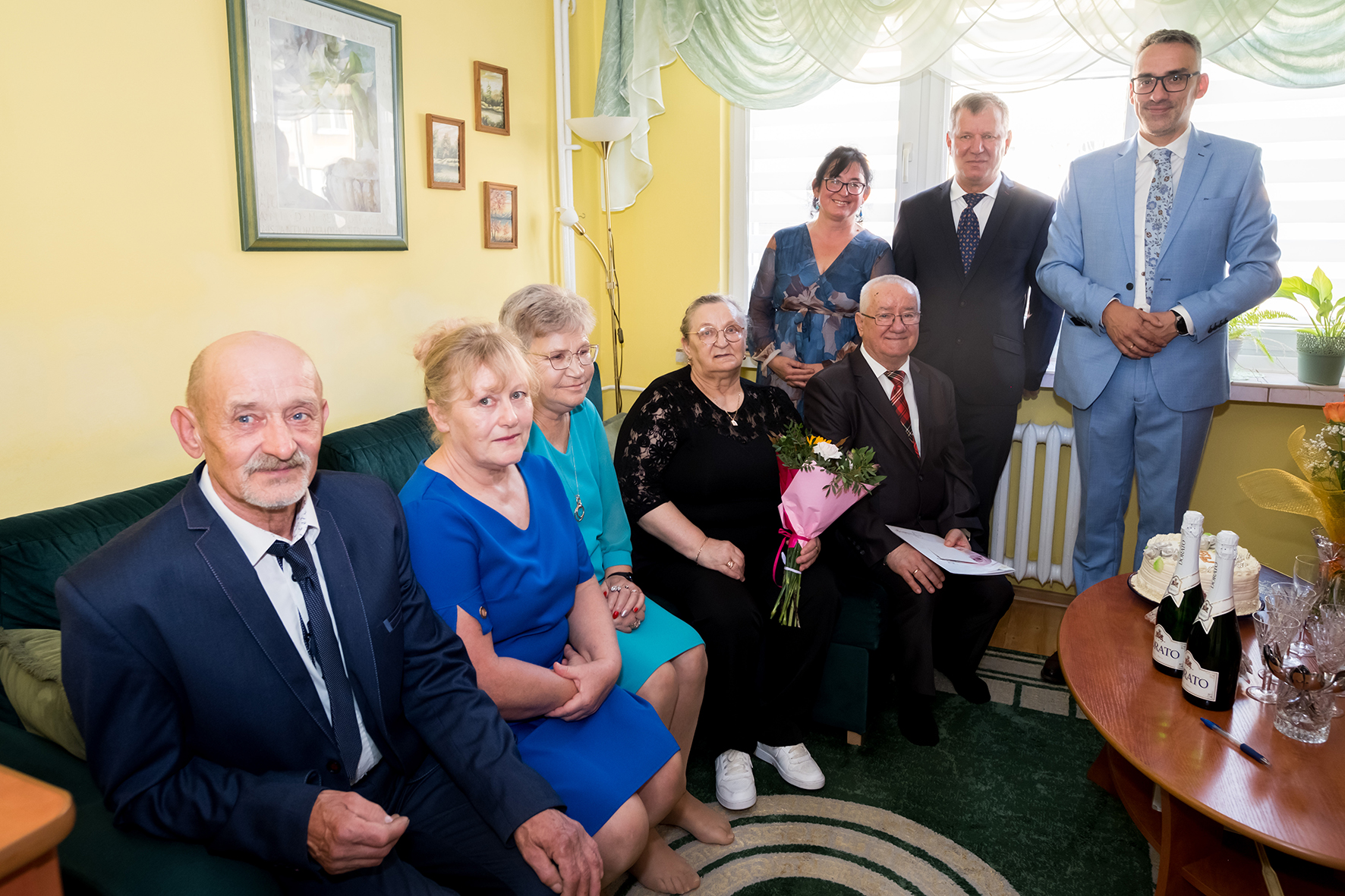 Jubilaci siedzą, obok inne osoby, na żółtej ścianie wiszą obrazki