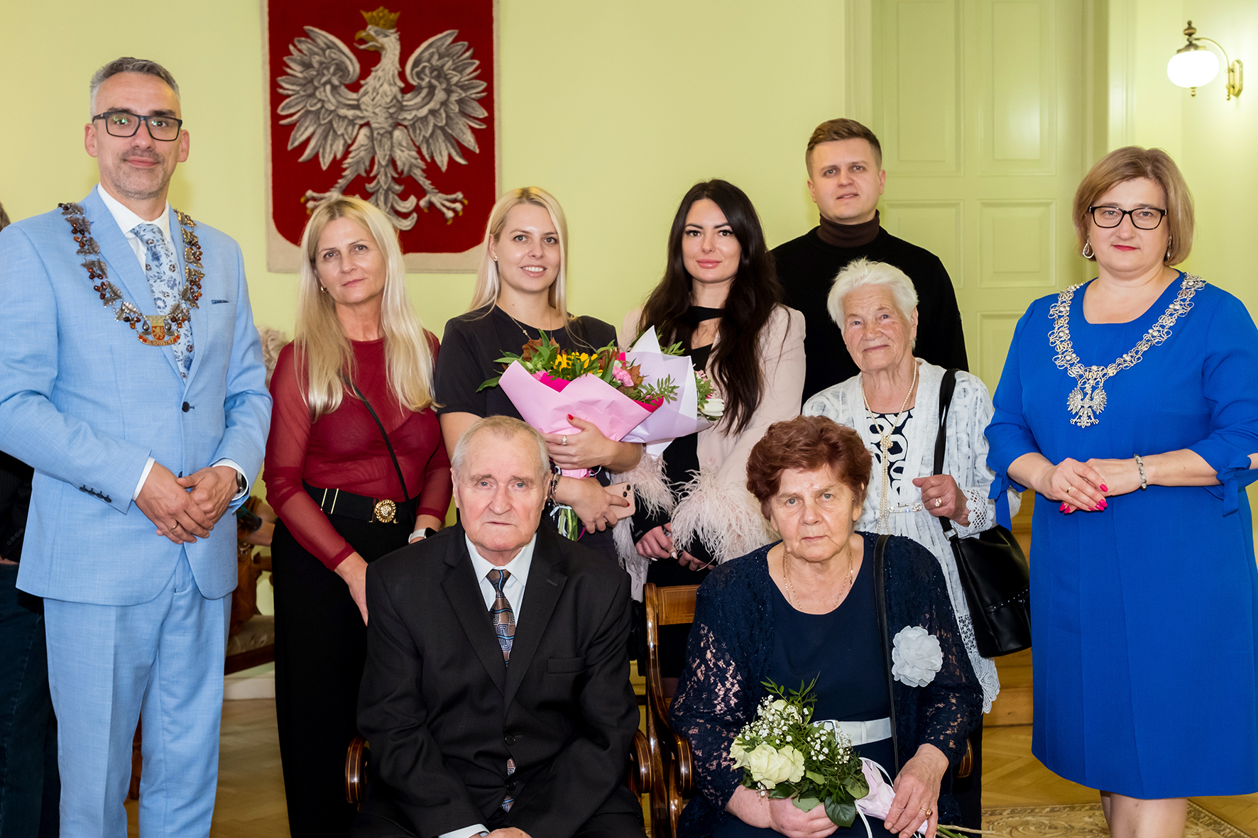 Jubilaci siedzą na krzesłach, za nimi stoja osoby z rodziny, w tle na ścianie wisi godło Polski
