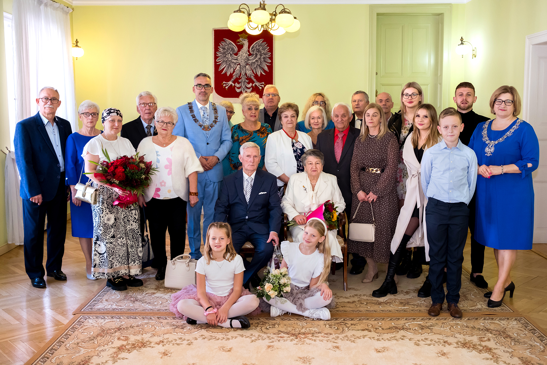 Jubilaci siedzą na krzesłach, za nimi stoja osoby z rodziny, w tle na ścianie wisi godło Polski