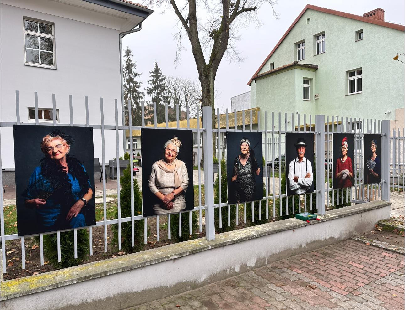 Wystawa portretów powieszona na płocie, w tle budynek 