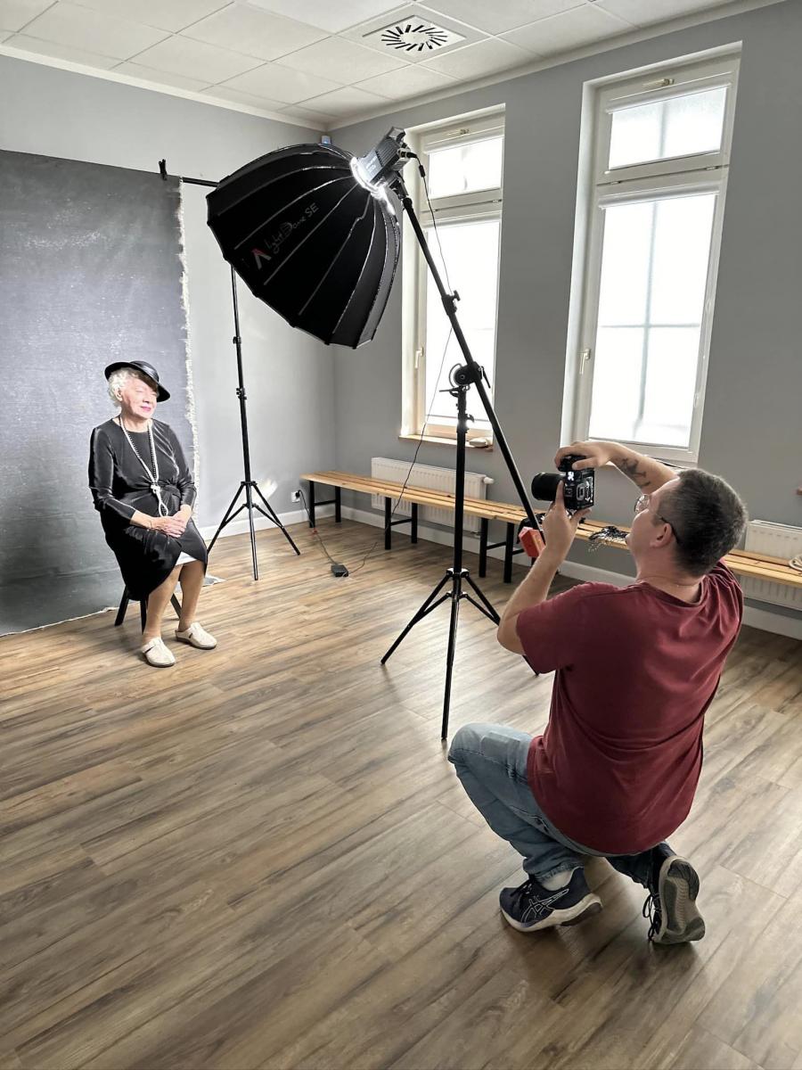 Praca studyjna. Starsza pani siedzi na krzesełku, w stylizacji oświetlenie, przed nią kuca fotograf z aparatem