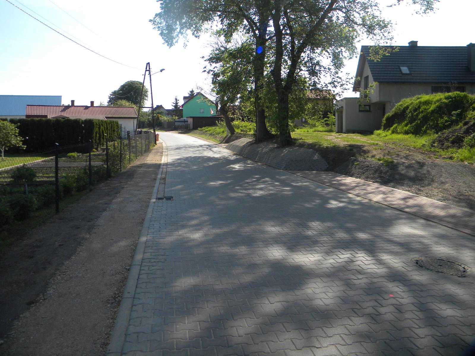 Jezdnia z szarej kostki polbruk, z lewej ogrodzenie z siatki, po prawej drzewa, w oddali budynki