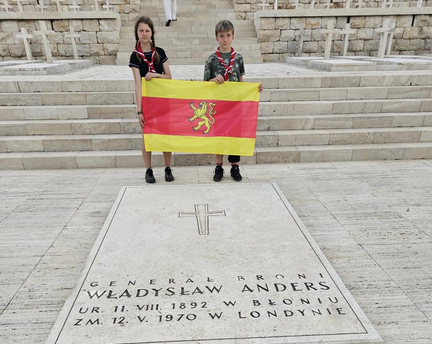 Dwóch harcerzy trzyma flagę Dębna na wzgórzu przed grobem Władysława Andersa