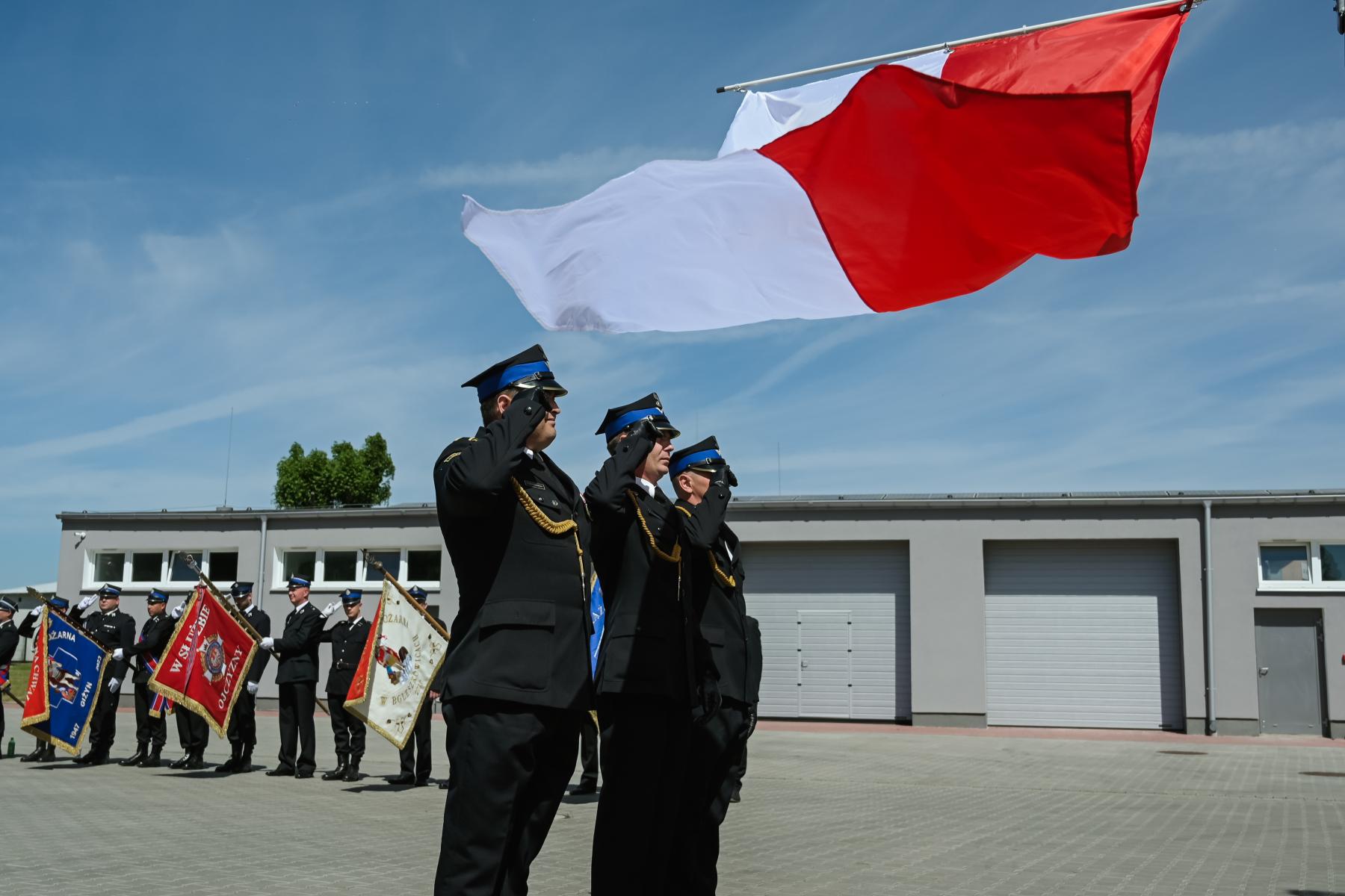 Strażacy w mundurach salutują. nad nimi powiewa biało czerwona flaga. Na placu, niebieskie niebo