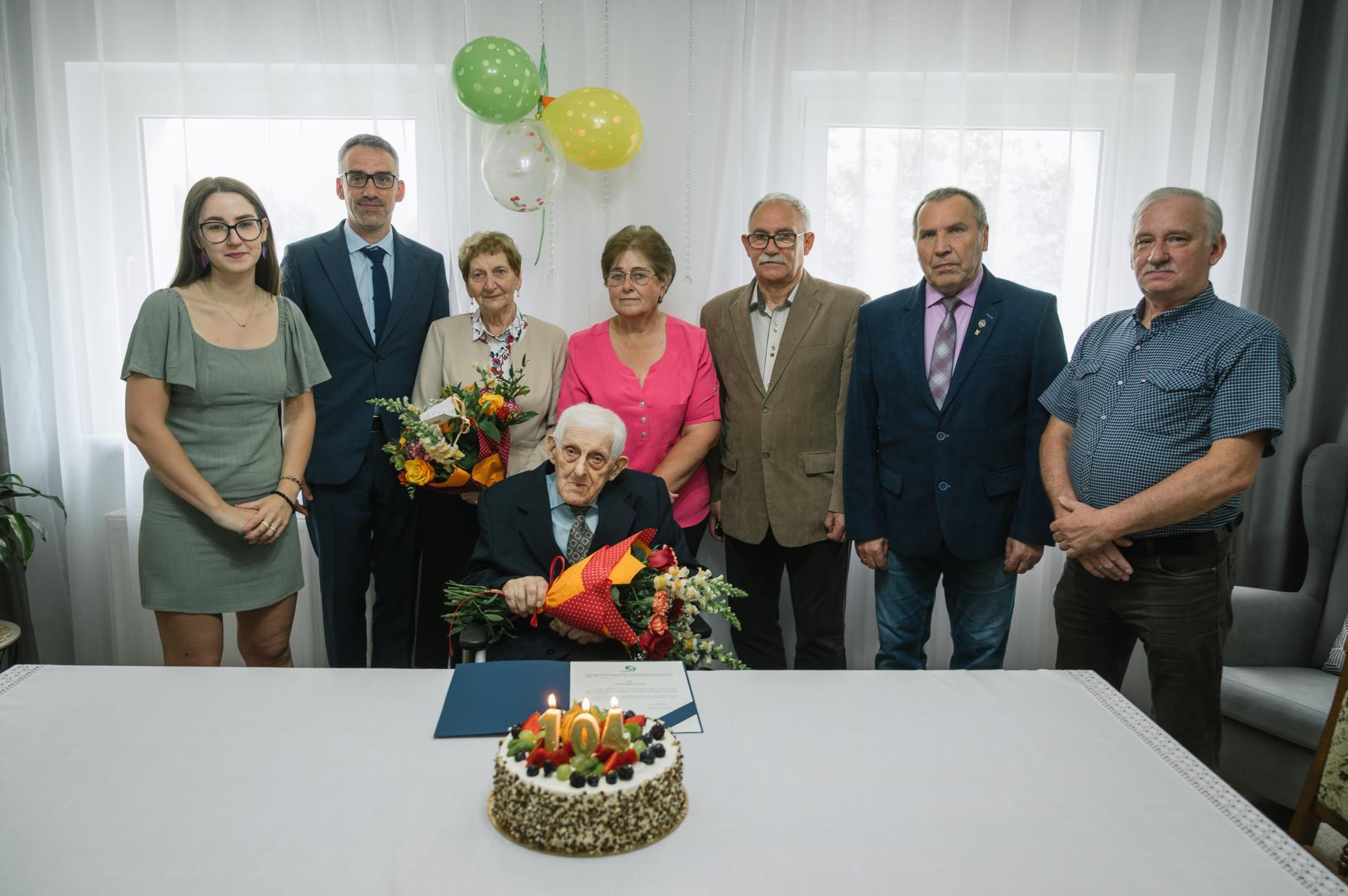 W pokoju stoją, od lewej: młoda kobieta, W. Czepułkowski, M. Mazur, kobieta, A. Kaczmarczyk, W. Sokalski, mężczyzna. przed nimi  siedzi Michał Markiewicz, trzyma w rękach bukiet kwiatów, na stole tort z palącymi świeczkami 104