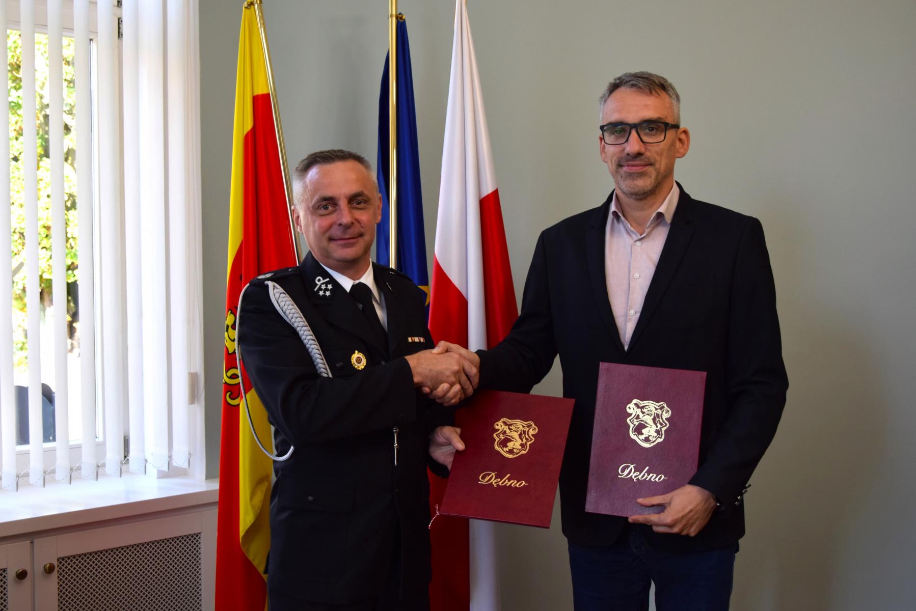 Od lewej, w gabinecie stoi w mundurze prezes OSP Krześnica, obok burmistrz Dębna, ściskają sobie dłoń w drugiej ręcę trzymają borodową teczkę. W tle flagi Polski, UE, Dębna
