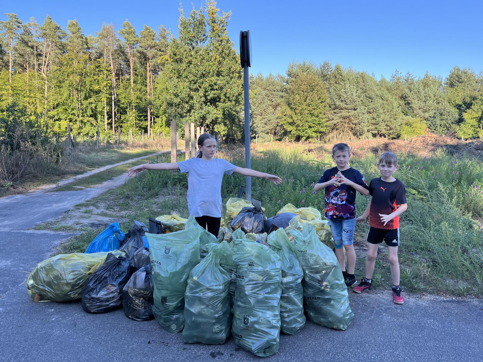 Dzieci stoją na końcu ściezki rowerowej. Przed nimi leżą worki ze śmieciami