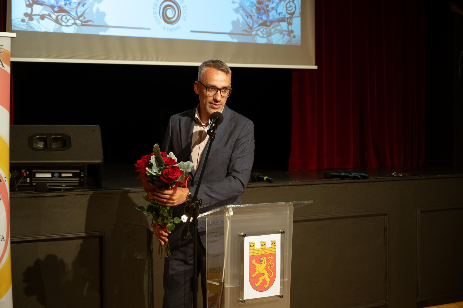 burmistrz Debna trzyma kwiaty w reku, stoi przy mównicy