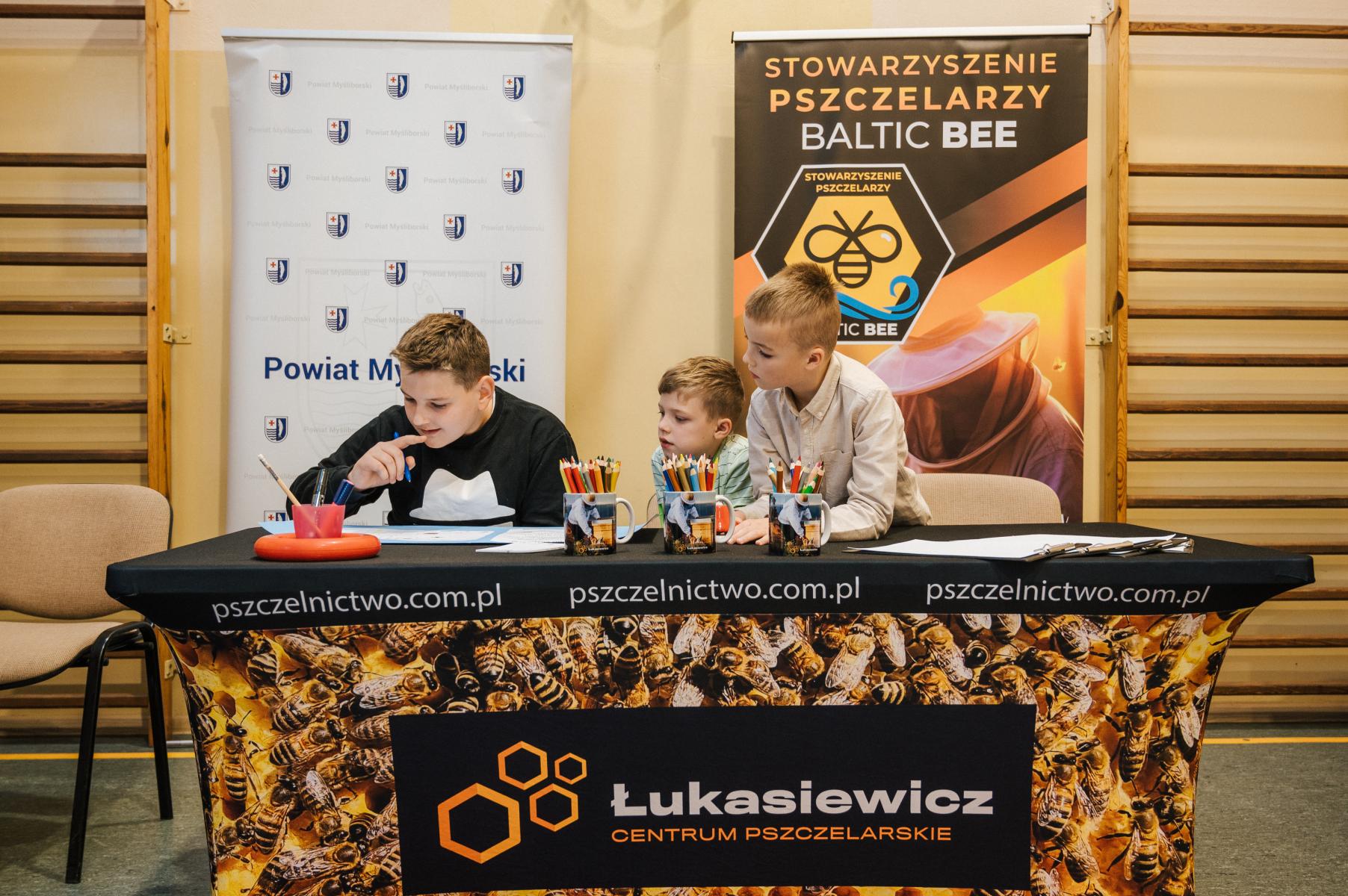 Trzech uczniów siedzą przy stole, wykonują jakieś zadanie, w tle banery