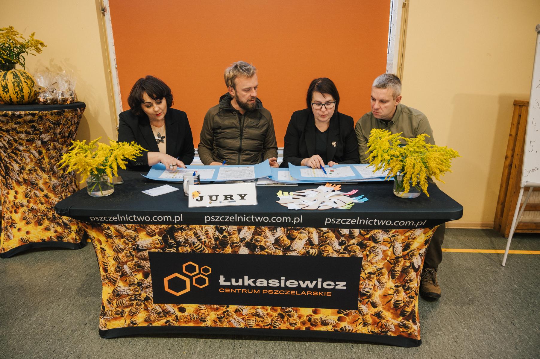 Czlonkowie jury, cztery osoby siedzą przy stole. Stół nakryty obrusem z pszczołami. 