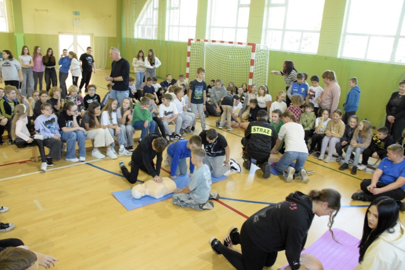 Duża grupa uczniów na hali. Cześć siedzi, część klęczy przed manekinami 